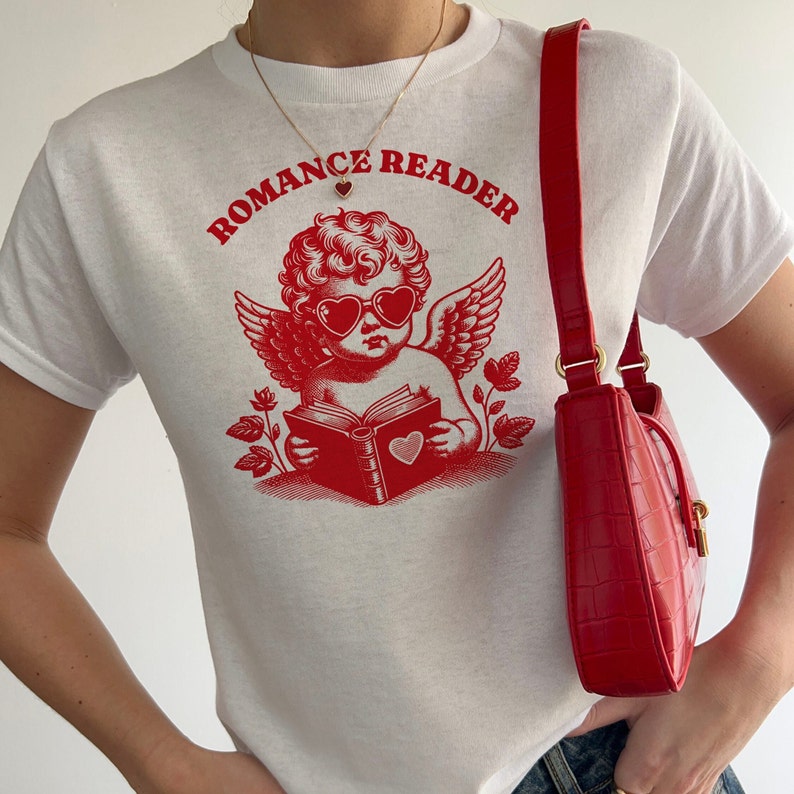 May include: White t-shirt with a red graphic of a cherub wearing heart-shaped sunglasses and reading a book. The text "Romance Reader" is printed above the cherub.