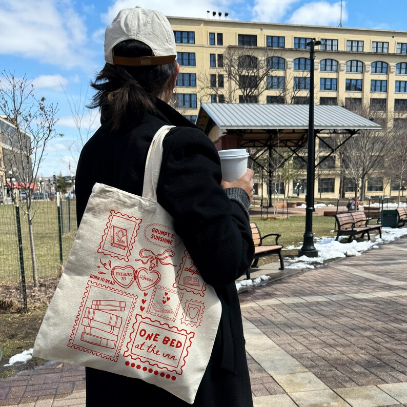 May include: A beige tote bag with a red stamp design featuring books, hearts, and the text "ONE BED at the inn." The bag is carried by a person wearing a black coat and a white cap, walking outdoors on a sunny day.