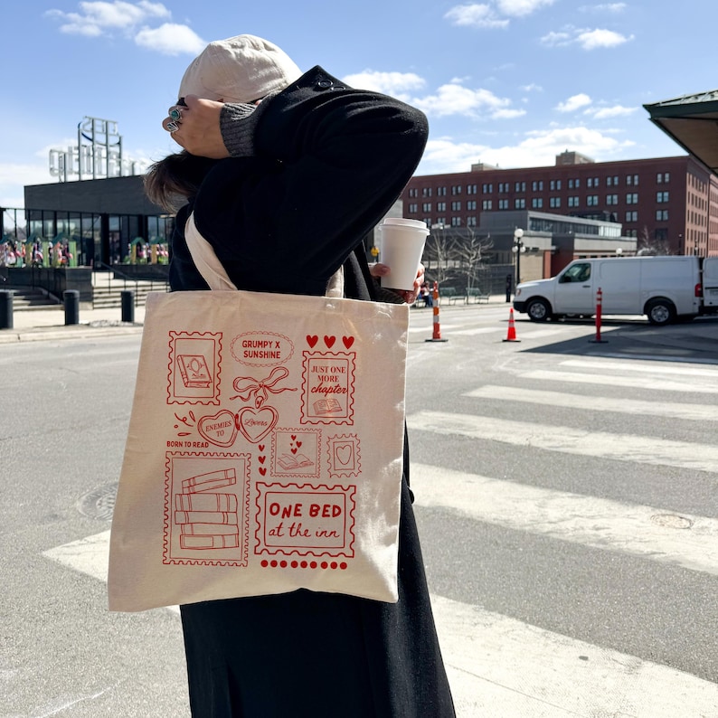 May include: A cream-colored tote bag with red illustrations and text. The design includes book stacks, hearts, and the phrases "Grumpy x Sunshine," "Just One More Chapter," and "One Bed at the Inn." The bag is carried by a person on a city street.