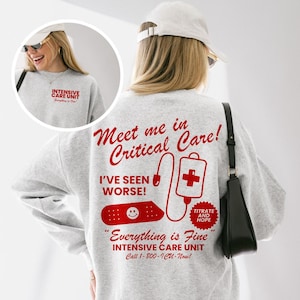 May include: A light gray sweatshirt with red text and graphics. The back of the sweatshirt says "Meet me in Critical Care!" with a medical theme. The front of the sweatshirt says "Intensive Care Unit" in red.