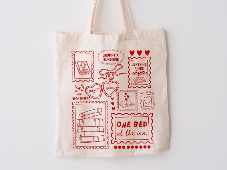 May include: A cream-colored tote bag featuring red illustrations of books, hearts, and stamps. Text includes "Born to Read," "Grumpy x Sunshine," and "One Bed at the Inn." The bag has a simple design with a focus on literary themes.