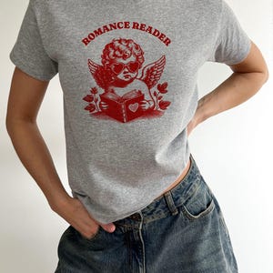 May include: A gray t-shirt with a red graphic of a cherub wearing heart-shaped sunglasses and holding a book with a heart on the cover. The text "ROMANCE READER" is printed above the cherub.