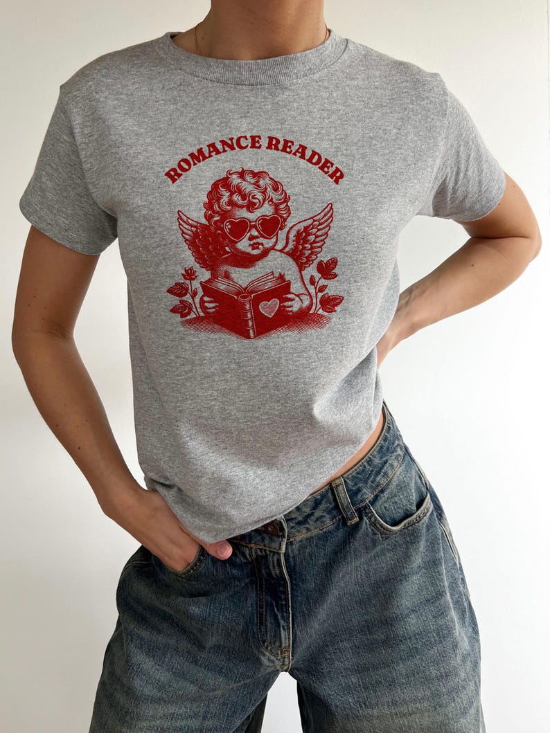 May include: A gray t-shirt with a red graphic of a cherub wearing heart-shaped sunglasses and holding a book with a heart on the cover. The text "ROMANCE READER" is printed above the cherub.