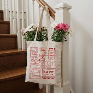 May include: A cream-colored tote bag with red graphic designs and text, including the phrases "Born to Read" and "One Bed at the Inn." The bag is filled with pink and white flowers. The tote bag is hanging on a white banister.