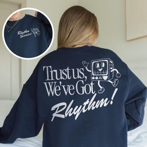 Op de afbeelding: Marineblauw sweatshirt met witte tekst en afbeeldingen. De achterkant heeft de zin "Trust us, We've Got Rhythm!" en een cartoon ECG-monitor karakter. De linkerborst heeft de tekst "Rhythm Specialist" en een bijpassende afbeelding.