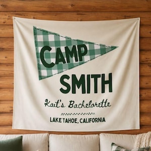 Pode incluir: Uma faixa creme com as palavras "CAMP SMITH" em verde escuro, acima de "Kait's Bachelorette" e "LAKE TAHOE, CALIFORNIA". No topo, há uma forma de flâmula xadrez verde e branca. A faixa está pendurada em uma parede de madeira.