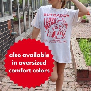 Puede incluir: Camiseta blanca con texto rojo que dice "But Daddy I Love Him" y una imagen de dibujos animados de una mujer con corazones rojos en las mejillas. El texto "24/7 Broken Hearts Club 365" tambi&eacute;n est&aacute; impreso en la camiseta.