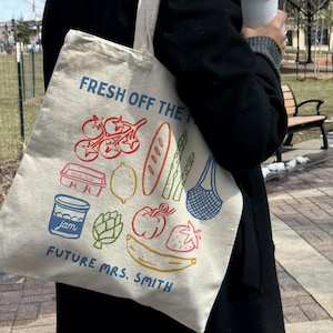 May include: A beige tote bag with colorful illustrations of food items like tomatoes, bread, and a banana. The text "FRESH OFF THE" and "FUTURE MRS. SMITH" are printed on the bag, which is being held outdoors.