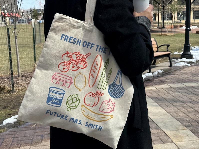 May include: A beige tote bag with colorful illustrations of food items like tomatoes, bread, and a banana. The text "FRESH OFF THE" and "FUTURE MRS. SMITH" are printed on the bag, which is being held outdoors.