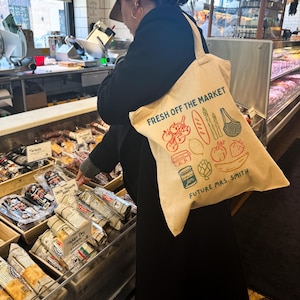 May include: Beige canvas tote bag with the text "FRESH OFF THE MARKET" and illustrations of produce. The bag also includes the text "FUTURE MRS. SMITH". The bag is being carried in a grocery store.