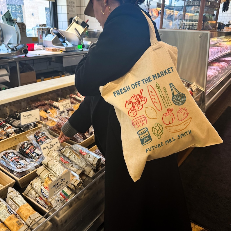 May include: Beige canvas tote bag with the text "FRESH OFF THE MARKET" and illustrations of produce. The bag also includes the text "FUTURE MRS. SMITH". The bag is being carried in a grocery store.