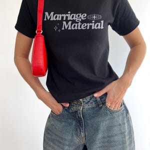 Puede incluir: Una camiseta negra con el texto "Marriage Material" en blanco con un peque&ntilde;o gr&aacute;fico de globo terr&aacute;queo y estrellas. La persona lleva pantalones vaqueros azules y un bolso de mano rojo.