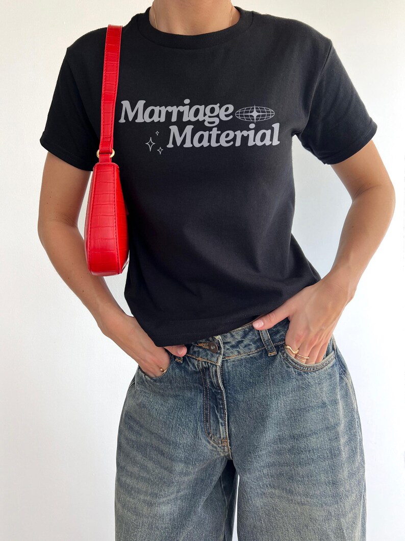 Puede incluir: Una camiseta negra con el texto "Marriage Material" en blanco con un peque&ntilde;o gr&aacute;fico de globo terr&aacute;queo y estrellas. La persona lleva pantalones vaqueros azules y un bolso de mano rojo.