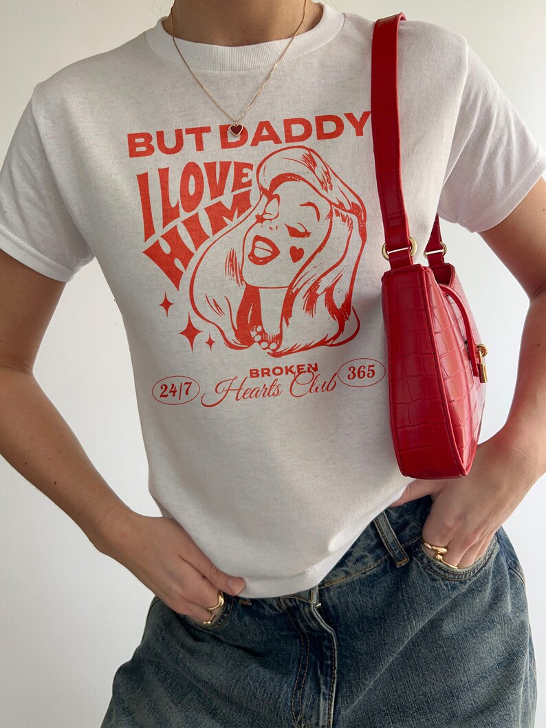 Puede incluir: Camiseta blanca con texto rojo que dice "But Daddy I Love Him" y una imagen de dibujos animados de una mujer con un coraz&oacute;n rojo en la mejilla. El texto tambi&eacute;n incluye "24/7 Broken Hearts Club 365".