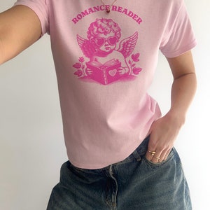 May include: Pink t-shirt with a graphic of a cherub wearing heart-shaped sunglasses and reading a book with a heart on the cover. The text "ROMANCE READER" is printed above the cherub.