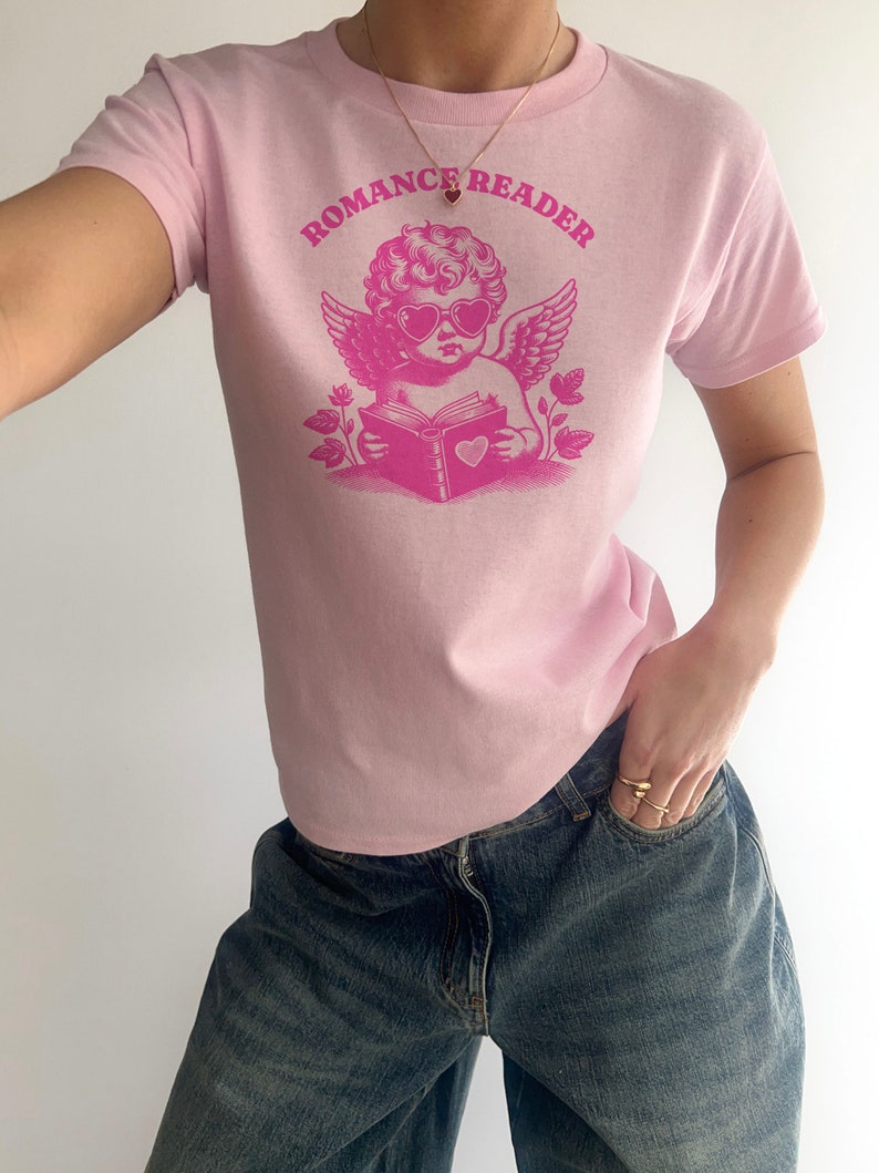 May include: Pink t-shirt with a graphic of a cherub wearing heart-shaped sunglasses and reading a book with a heart on the cover. The text "ROMANCE READER" is printed above the cherub.