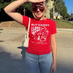 Puede incluir: Una camiseta roja con un estampado gr&aacute;fico blanco que dice "But Daddy I Love Him" y "24/7 Broken Hearts Club 365".