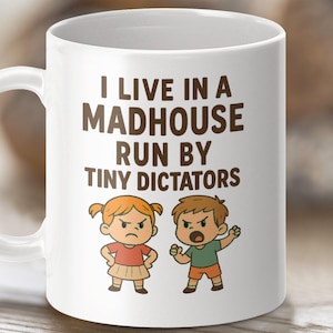 以下が含まれることがあります： 白いコーヒーカップに、茶色の文字で「私は小さな独裁者によって運営されている精神病院に住んでいます」と書かれています。カップには、赤い髪の女の子と茶色の髪の男の子の2人の子供が描かれており、どちらも怒った表情をしています。
