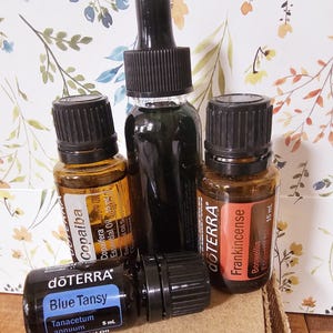 May include: Four amber glass bottles of essential oils with black dropper tops. The bottles are labeled with the brand name "dōTERRA" and the names of the oils: "Copaiba", "Blue Tansy", "Frankincense", and "Blue Tansy". The bottles are sitting on a brown cardboard surface.
