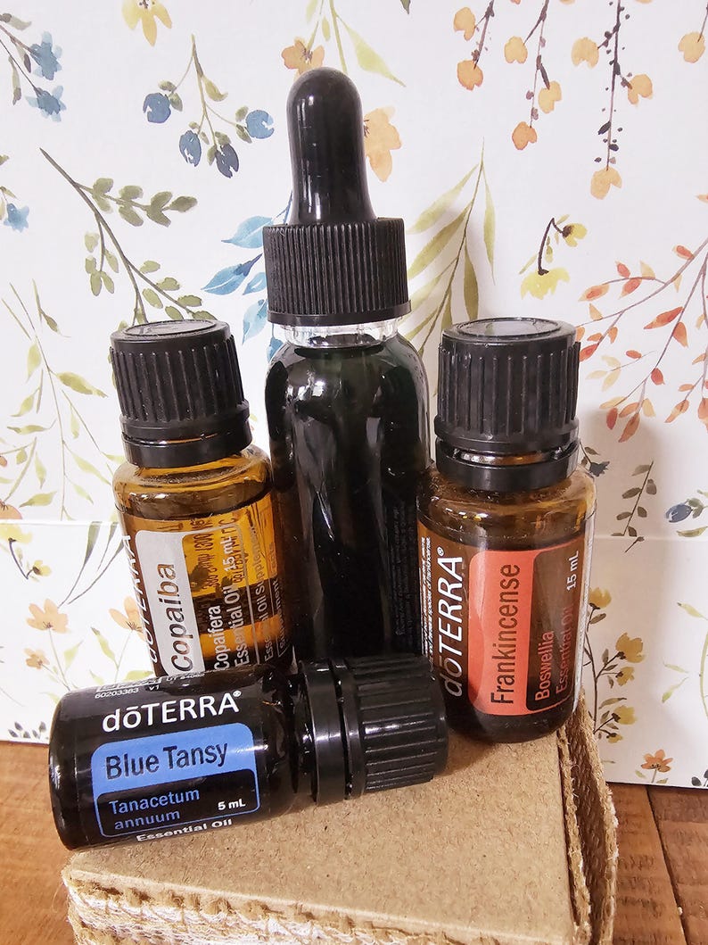May include: Four amber glass bottles of essential oils with black dropper tops. The bottles are labeled with the brand name "dōTERRA" and the names of the oils: "Copaiba", "Blue Tansy", "Frankincense", and "Blue Tansy". The bottles are sitting on a brown cardboard surface.