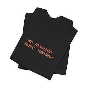 May include: Black t-shirt with the text "WE REQUIRE MORE TOKENS!" in orange pixelated font. The shirt is folded, showing the neckline and part of the front. The text is in a retro video game style.