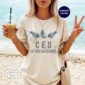 May include: Ivory-colored t-shirt with a graphic of three gray pigeons and the text "CEO OF BREADCRUMBS." The shirt also has the words "COMFORT COLORS" and "COLOR IVORY" printed on it. The shirt is a casual wear item.