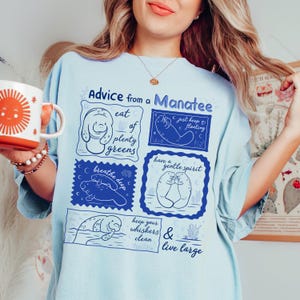May include: Light blue t-shirt with the text "Advice from a Manatee" and illustrations of manatees with phrases like "eat plenty greens" and "live large". The shirt is a relaxed fit.