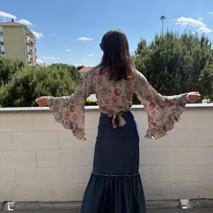 May include: A floral print blouse with long, flared sleeves and a denim maxi skirt. The blouse has a tie-back detail. The skirt features a ruffled hem. The outfit is worn outdoors on a sunny day.