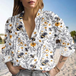 May include: White button-down shirt with a repeating pattern of bumblebees, yellow flowers, and gray foliage. The shirt has long sleeves and a V-neck collar. The background is a blurred beach scene.