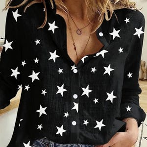 Pode incluir: Camisa preta com estampa de estrelas brancas. A camisa de manga comprida tem uma gola clássica e está parcialmente desabotoada. A camisa é combinada com jeans azul.