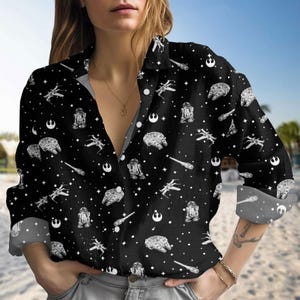 Puede incluir: Camisa negra con botones con un estampado de Star Wars. La camisa presenta ilustraciones blancas de naves espaciales, droides y símbolos de estrellas dispersos por la tela. Las mangas largas están dobladas, revelando un interior gris.