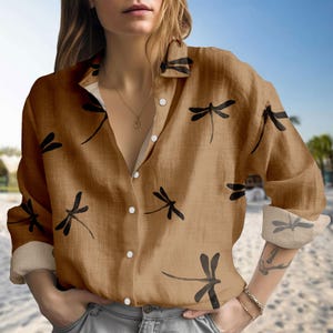 May include: A tan button-down shirt featuring a black dragonfly design. The long-sleeved shirt has a classic collar and cuffs, with white buttons. The fabric appears to be a lightweight material. The shirt is suitable for casual wear.