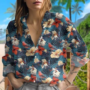 Può includere: Una camicia button-down color verde acqua con un vivace motivo floreale e di uccelli. Il design presenta fiori rossi, crema e gialli, insieme a uccelli rossi, su uno sfondo di foglie verde scuro. La camicia ha maniche lunghe e un colletto classico.