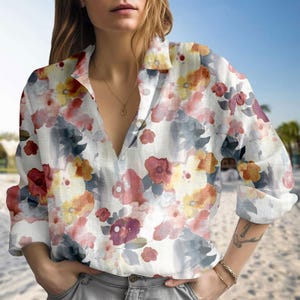 Puede incluir: Camisa blanca abotonada con un estampado floral de acuarela en tonos rojos, amarillos y azules. La camisa de manga larga tiene un cuello clásico y está desabrochada en el escote.