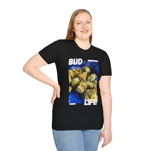 Puede incluir: Camiseta negra con un diseño rectangular que muestra cogollos verdes y amarillos sobre un fondo azul, con las palabras "BUD LIFE" en blanco. La camiseta es de cuello redondo.
