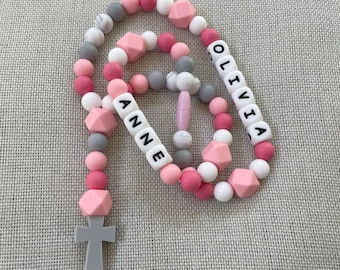 May include: A beaded necklace featuring a mix of round and geometric silicone beads in shades of pink, white, and gray. The necklace includes white letter beads spelling out the names ANNE and OLIVIA, and it terminates with a gray silicone cross pendant.