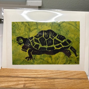 May include: A rectangular art print featuring a black silhouette of a turtle against a textured, mottled lime green and yellow background. The artwork is signed with the initials RH in the bottom right corner and is enclosed in a clear sleeve.