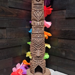May include: A brown wooden tiki statue with four carved faces and a base. The statue is decorated with a colorful lei.