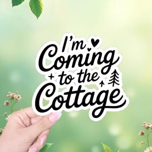 May include: A white sticker with black cursive text that reads "I'm Coming + to the Cottage". The design includes a black heart, a plus sign, a small tree, and an upward arrow. The sticker is held by a hand with a blurred green background.