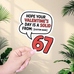 May include: A white sticker with a black outline, featuring the text "HOPE YOUR VALENTINE'S DAY IS A SOLID FROM: [CUSTOM NAME]" in a speech bubble. Below is the number "67" in red with a black outline.