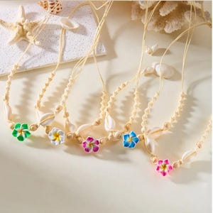 May include: Five necklaces with cream-coloured cords, each adorned with a seashell and a colourful flower pendant in green, white, purple, blue, and pink. The necklaces are displayed on a light-coloured surface.