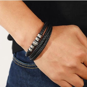 May include: Black braided leather bracelet with silver-tone metal accents. The bracelet wraps around the wrist twice, featuring a woven design and decorative metal beads. A stylish accessory for everyday wear.
