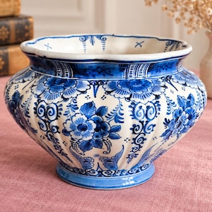 Pode incluir: Um vaso de cerâmica com base branca e padrões florais azuis complexos e desenhos em volutas. O recipiente tem uma borda larga e recortada, um corpo arredondado com detalhes estriados verticais e uma base circular pintada à mão.