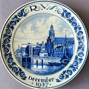 Może przedstawiać: Ozdobny ceramiczny talerz w kolorze niebiesko-białym. Talerz przedstawia szczegółową ilustrację sceny miejskiej nad wodą, z kościołem i łodziami. Obrzeże zdobią wzory kwiatowe, a widoczny jest napis "Grudzień 1937".