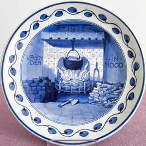 May include: A blue and white ceramic plate with a Dutch scene depicting a fireplace with a pot hanging over the fire. The text "Vrienden in Nood" and "A 1918" are visible on the plate.