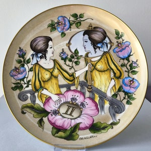 Op de afbeelding: Een decoratief keramiek bord met een gouden rand heeft een handgeschilderd ontwerp van twee vrouwen in gele jurken, een houdt een spiegel vast, omringd door bloemen en het Romeinse cijfer II. Het bord is gesigneerd met "Ole Winther".