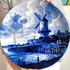Peut inclure: Une assiette en céramique bleue et blanche avec une scène peinte d'un moulin à vent dans un paysage hollandais. L'assiette représente un moulin à vent avec une voile blanche, une rivière et un ciel nuageux.