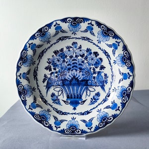 Könnte beinhalten: Ein weißer Teller aus Keramik mit einem blauen und weißen Blumendesign. Der Teller zeigt einen Blumenkorb mit zwei Vögeln auf jeder Seite. Der Teller hat einen gewellten Rand.