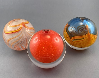 May include: Three glass spheres with different patterns. The first sphere is white with orange and yellow stripes. The second sphere is orange with red speckles. The third sphere is blue with orange and yellow stripes.