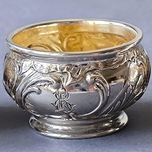 Peut inclure: Un petit saleron rond en métal argenté avec un intérieur doré. L'extérieur présente un motif ornemental en relief avec des volutes et un monogramme central gravé composé des lettres R et O entrelacées dans une police cursive.